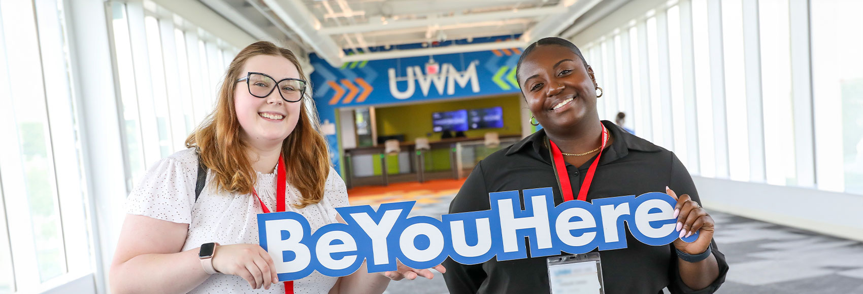 Two UWM team members smiling and holding a sign that says Be You Here